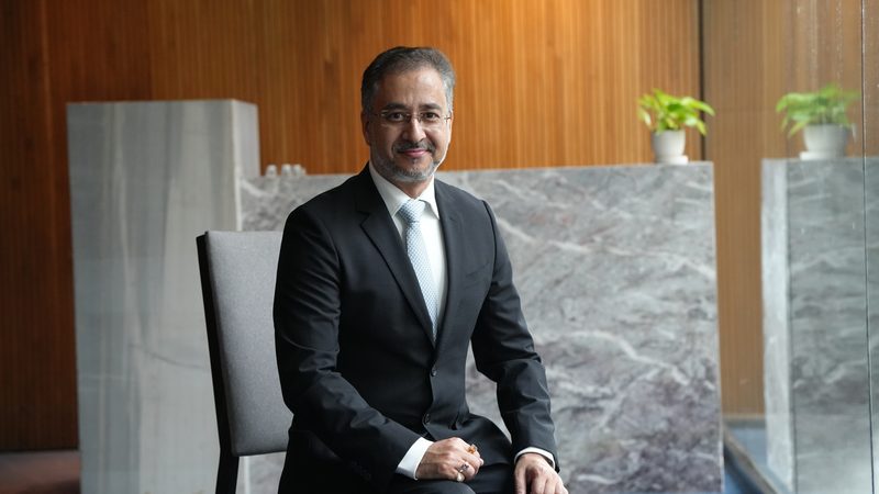 Avinash Kaul seated in a dark suit against a wood-panelled wall, smiling at the camera