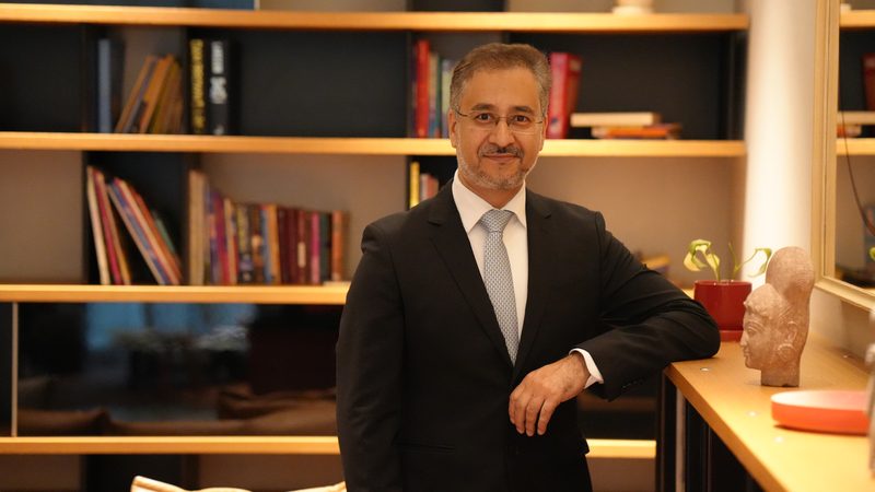 Avinash Kaul in a dark suit leaning on a counter in front of bookshelves