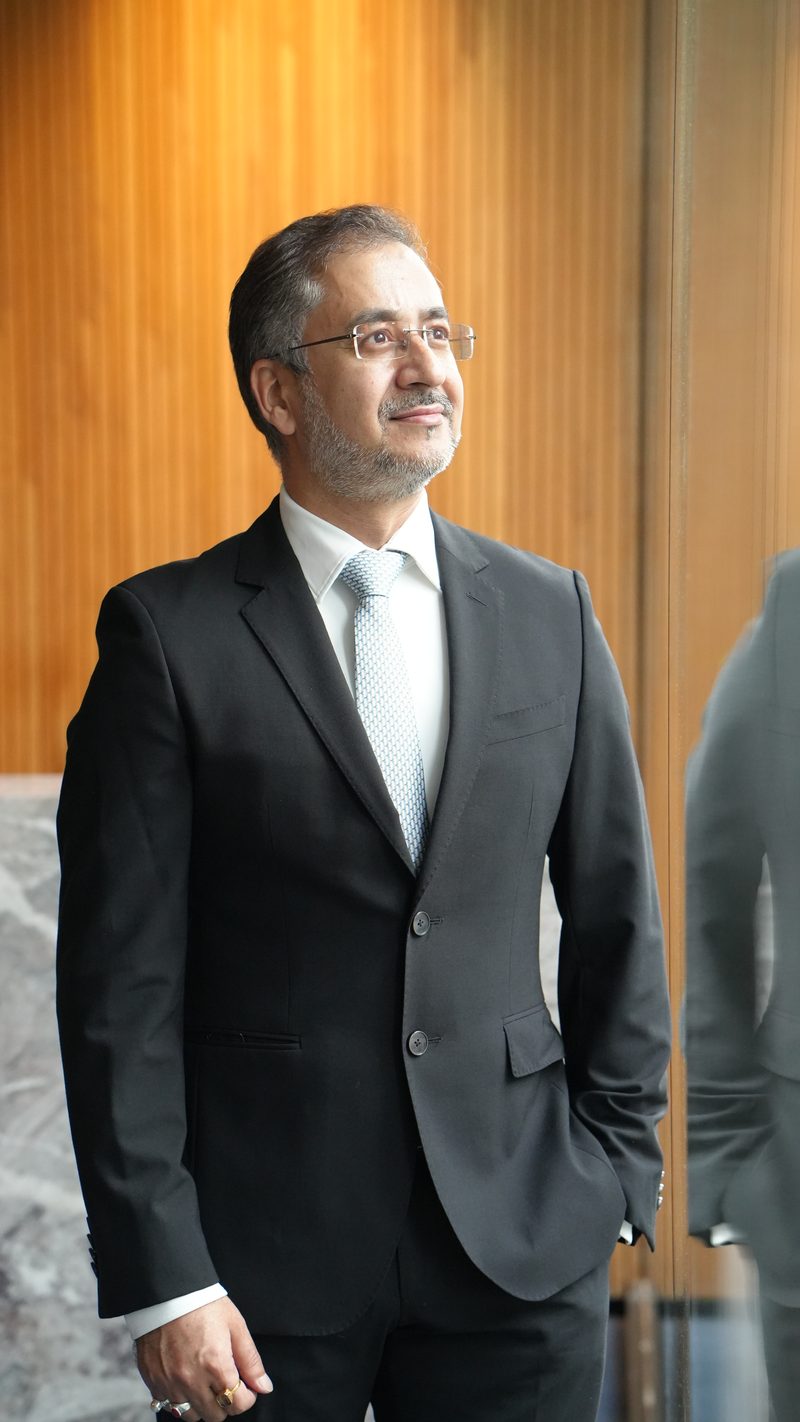 Avinash Kaul in a dark suit, hand in pocket, looking away from the camera against a wooden wall