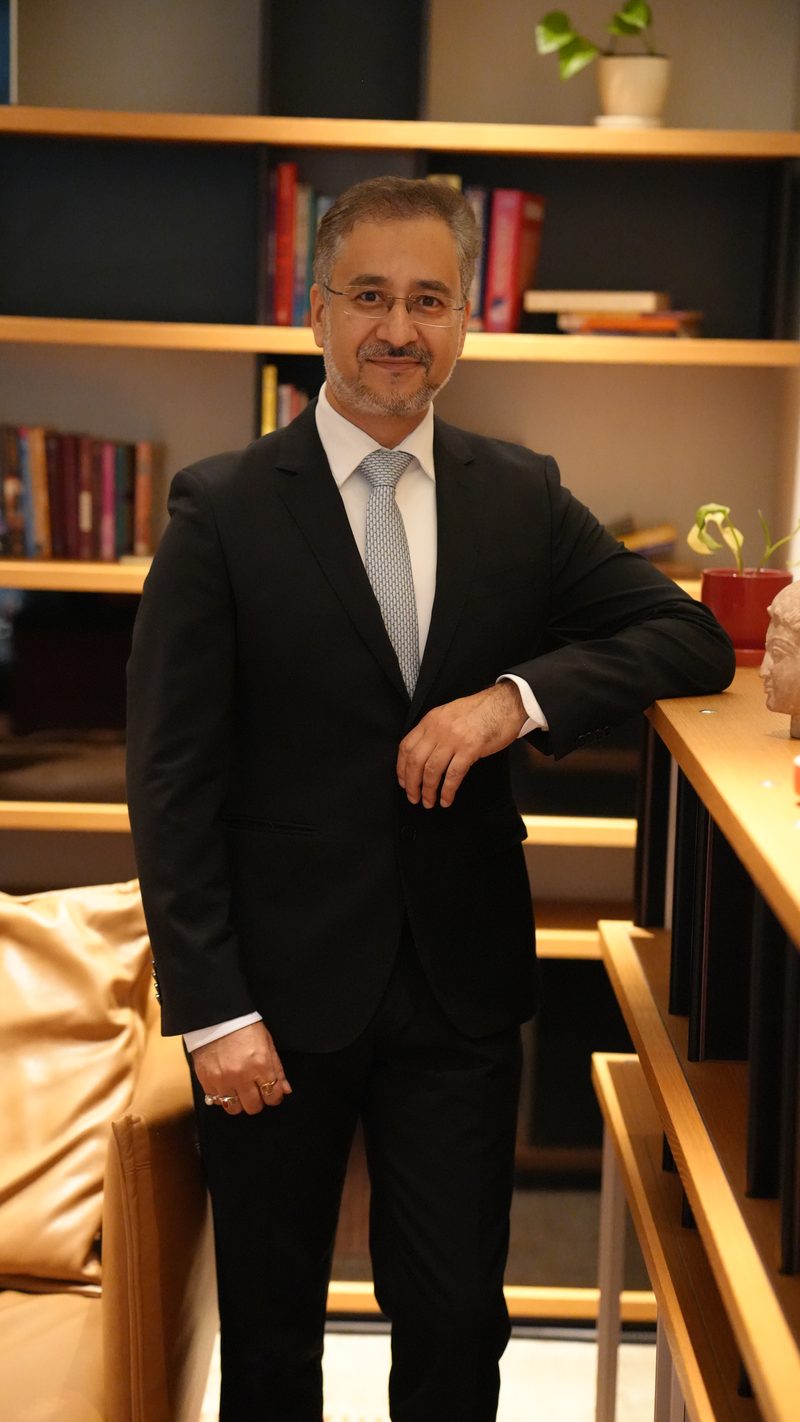 Avinash Kaul in a dark suit leaning on a shelf in a library setting, smiling