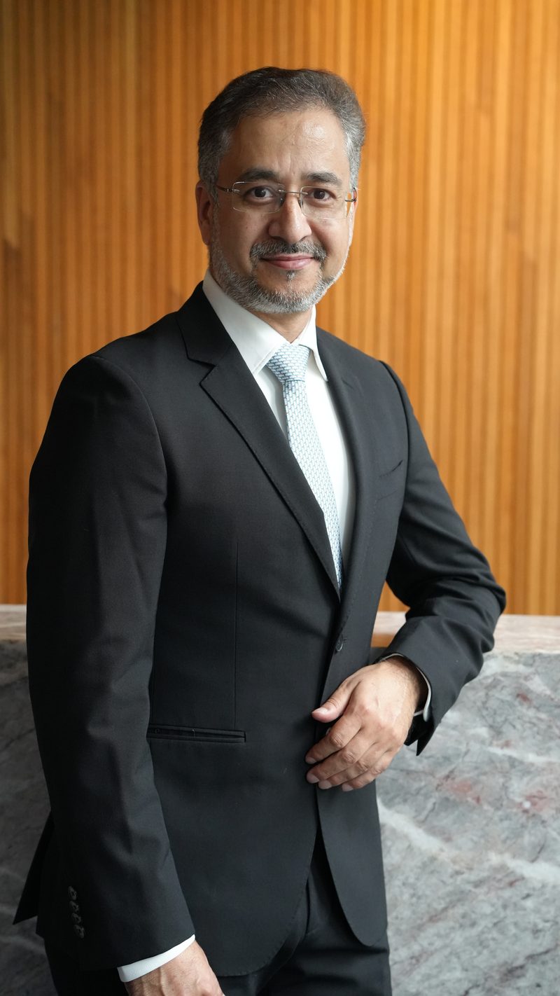 Avinash Kaul in a dark suit, standing with one hand at his waist, smiling at the camera